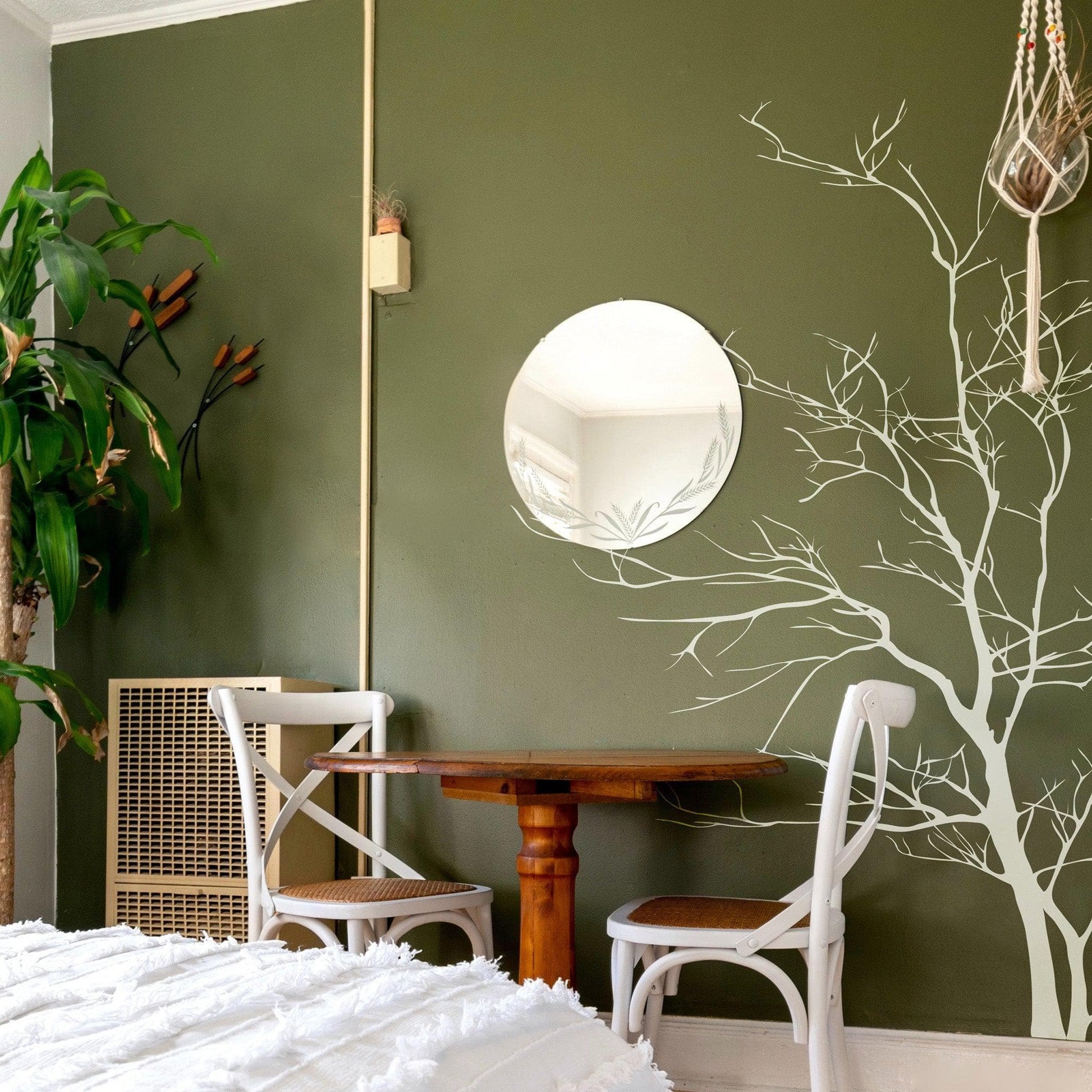 A white tree decal on a dark green wall in a bedroom above a brown table and two chairs, near a circular  mirror.