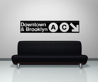 A black decal on a white wall showing "Downtown and Brooklyn" with the letters A and C in circular shapes and a white arrow pointing to the bottom right corner of the decal. Under it is a black couch.