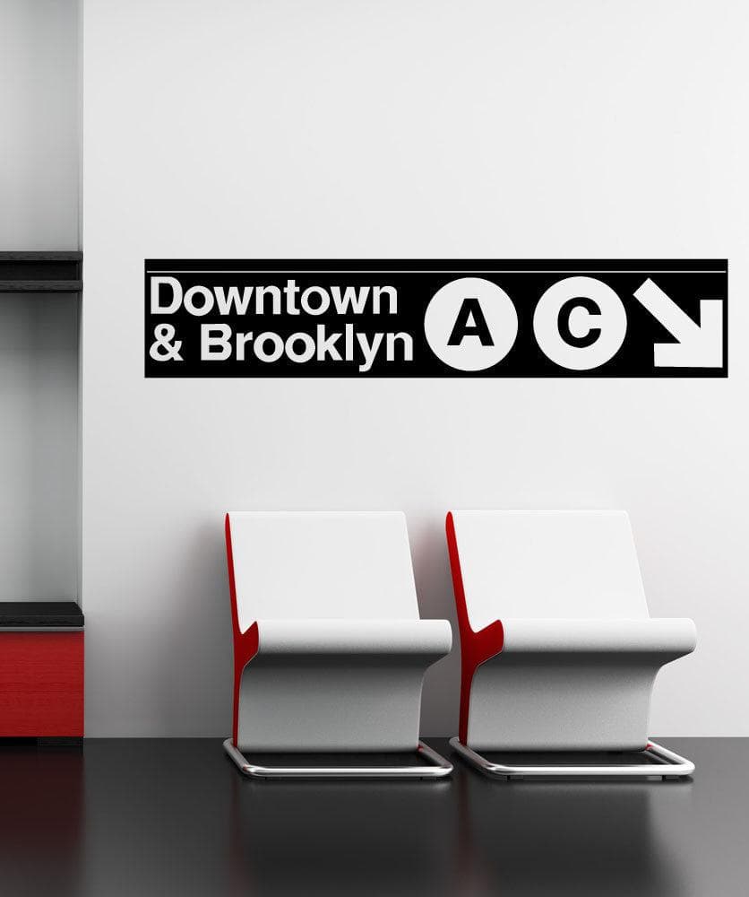 A black decal on a white wall showing "Downtown and Brooklyn" with the letters A and C in circular shapes and a white arrow pointing to the bottom right corner of the decal. Under it are two white chairs.