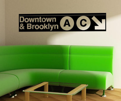 A black decal on a white wall showing "Downtown and Brooklyn" with the letters A and C in circular shapes and a white arrow pointing to the bottom right corner of the decal. Under it is a green couch.