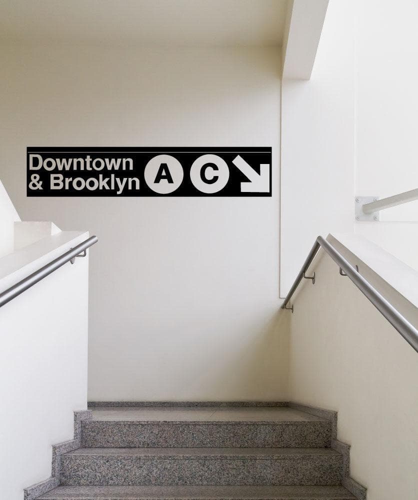 A black decal on a white wall showing "Downtown and Brooklyn" with the letters A and C in circular shapes and a white arrow pointing to the bottom right corner of the decal. Under it is a gray staircase.