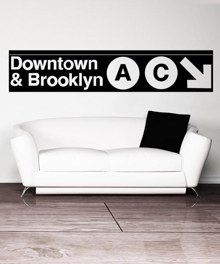 A black decal on a white wall showing "Downtown and Brooklyn" with the letters A and C in circular shapes and a white arrow pointing to the bottom right corner of the decal. Under it is a white couch with a black pillow.