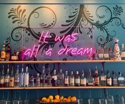 A large black floral swirl decal on a blue wall above a shelf of bottles, with a neon sign underneath it saying: "It was all a dream."