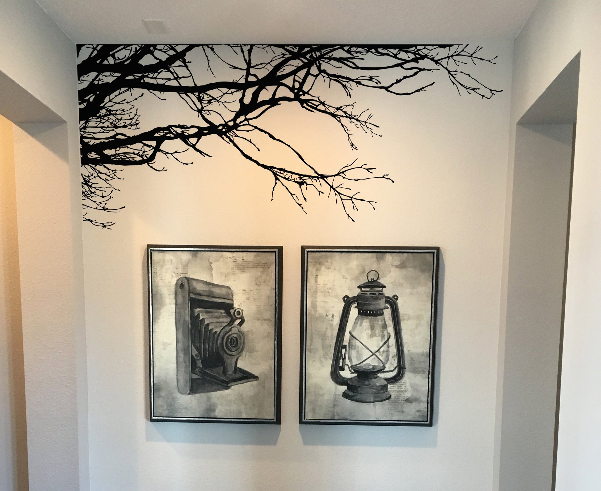 A white wall with a black tree decal, with framed photographs of an old camera and a lantern underneath it.