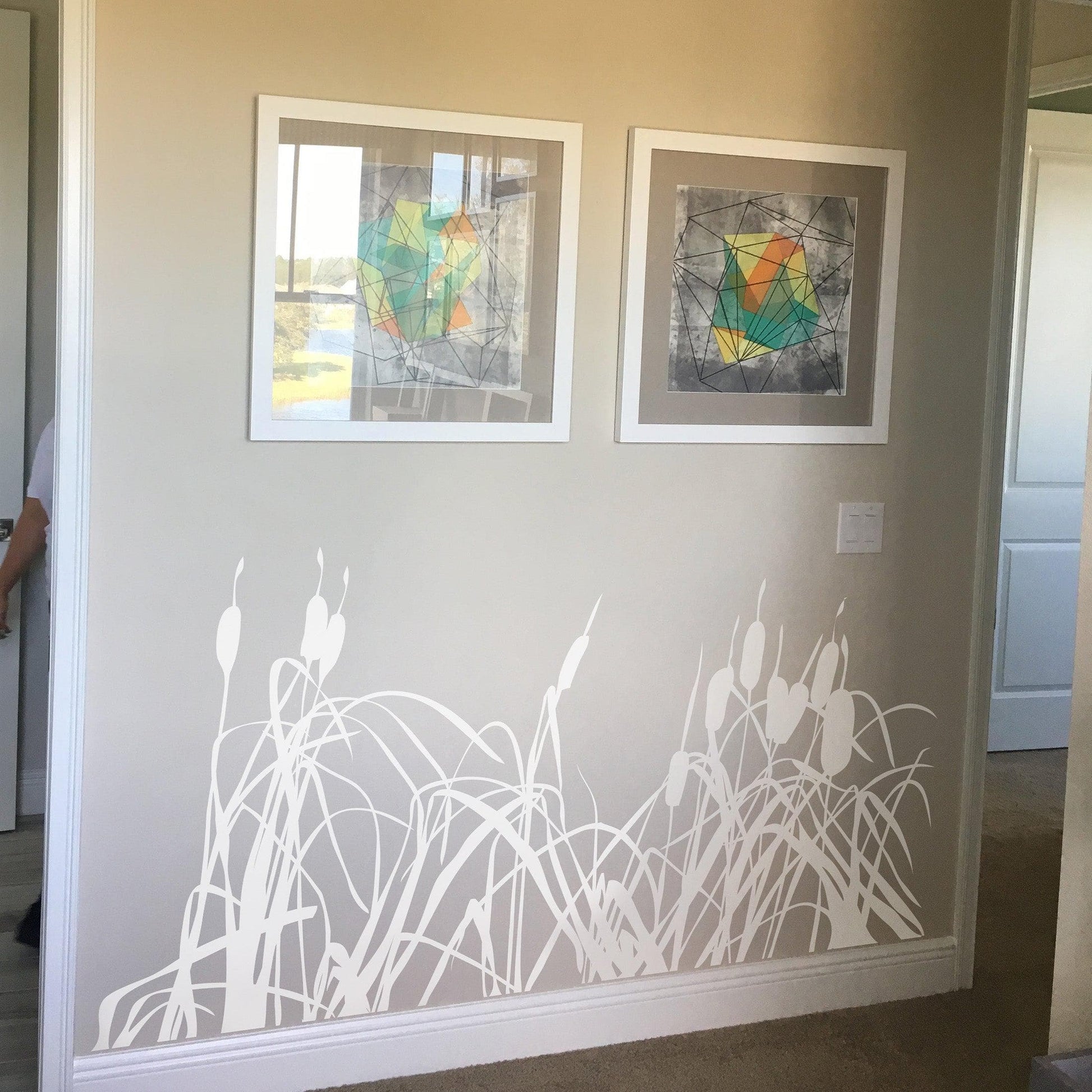 A white cattails decal on a white wall below two white framed photographs.