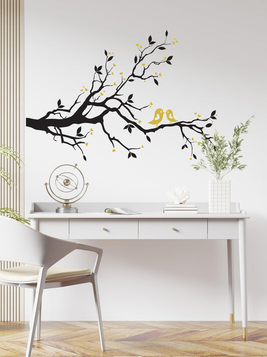 A black decal of a tree branch with two birds on a white wall above a desk.